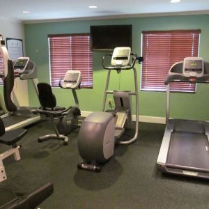 The hotel fitness center has two windows fitted with blinds, a wall-mounted TV, and  assorted exercise machines.