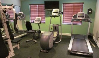 The hotel fitness center has two windows fitted with blinds, a wall-mounted TV, and  assorted exercise machines.