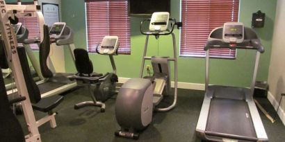 The hotel fitness center has two windows fitted with blinds, a wall-mounted TV, and  assorted exercise machines.