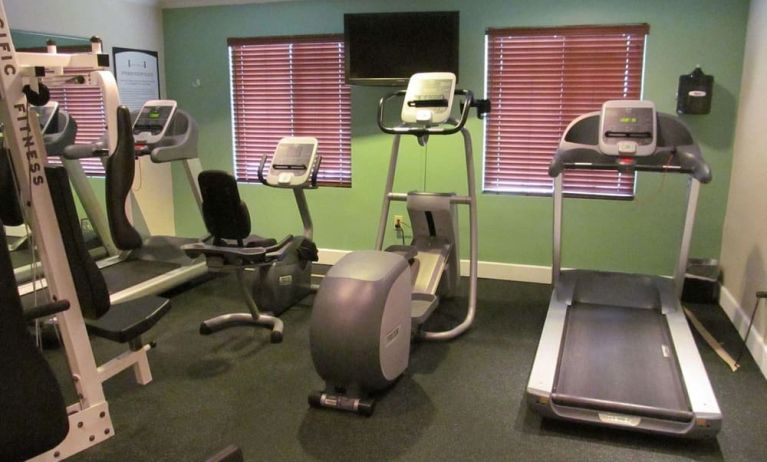 The hotel fitness center has two windows fitted with blinds, a wall-mounted TV, and  assorted exercise machines.