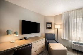 Sonesta Simply Suites Jersey City guest room workspace, including desk, chair, and lamp, with nearby TV and armchair.