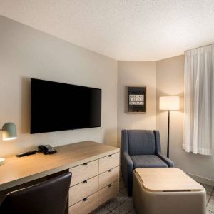 Sonesta Simply Suites Jersey City guest room workspace, including desk, chair, and lamp, with nearby TV and armchair.