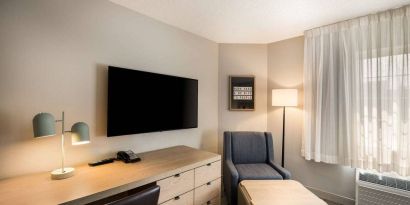 Sonesta Simply Suites Jersey City guest room workspace, including desk, chair, and lamp, with nearby TV and armchair.