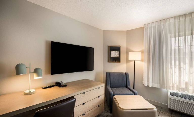 Sonesta Simply Suites Jersey City guest room workspace, including desk, chair, and lamp, with nearby TV and armchair.