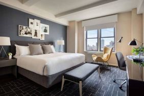 Double bed guest room in The Shelburne Sonesta New York, featuring a window, art on the wall, and a workspace desk, chair, and lamp.