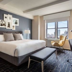 Double bed guest room in The Shelburne Sonesta New York, featuring a window, art on the wall, and a workspace desk, chair, and lamp.