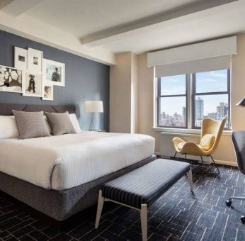 Double bed guest room in The Shelburne Sonesta New York, featuring a window, art on the wall, and a workspace desk, chair, and lamp.