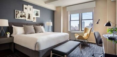 Double bed guest room in The Shelburne Sonesta New York, featuring a window, art on the wall, and a workspace desk, chair, and lamp.