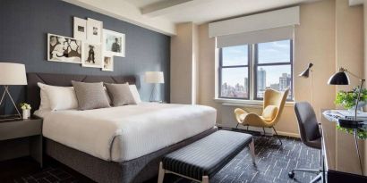 Double bed guest room in The Shelburne Sonesta New York, featuring a window, art on the wall, and a workspace desk, chair, and lamp.