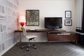 Guest room workspace in The Fifty Sonesta Select New York, featuring desk, chair, and lamp, with art on the wall and a nearby TV.