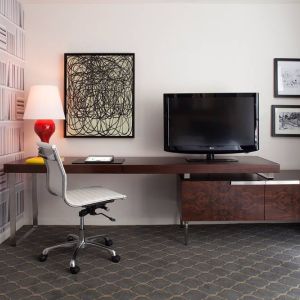 Guest room workspace in The Fifty Sonesta Select New York, featuring desk, chair, and lamp, with art on the wall and a nearby TV.