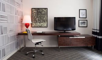 Guest room workspace in The Fifty Sonesta Select New York, featuring desk, chair, and lamp, with art on the wall and a nearby TV.