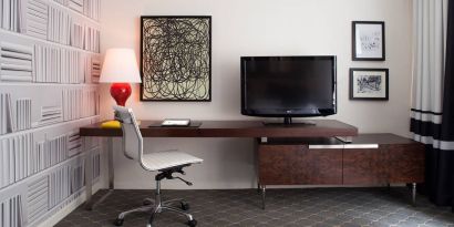 Guest room workspace in The Fifty Sonesta Select New York, featuring desk, chair, and lamp, with art on the wall and a nearby TV.