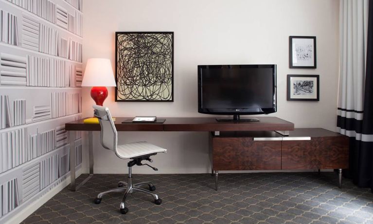 Guest room workspace in The Fifty Sonesta Select New York, featuring desk, chair, and lamp, with art on the wall and a nearby TV.