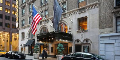 The Benjamin Royal Sonesta New York’s exterior features the hotel’s name on the awning and flag, with a Stars and Stripes flag beside it, and large potted plants.