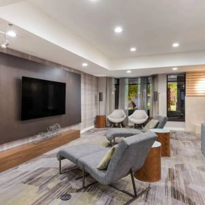 The hotel lobby includes a large, wall-mounted TV faced by a pair of reclined seats, with a fireplace and sofa nearby.