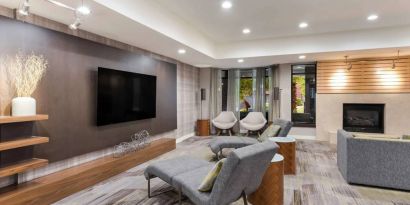 The hotel lobby includes a large, wall-mounted TV faced by a pair of reclined seats, with a fireplace and sofa nearby.