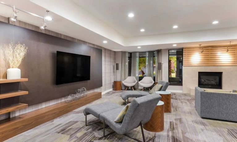 The hotel lobby includes a large, wall-mounted TV faced by a pair of reclined seats, with a fireplace and sofa nearby.