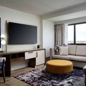 Sonesta Irvine guest room living area, including corner sofa, chair, window, and large, wall-mounted TV.