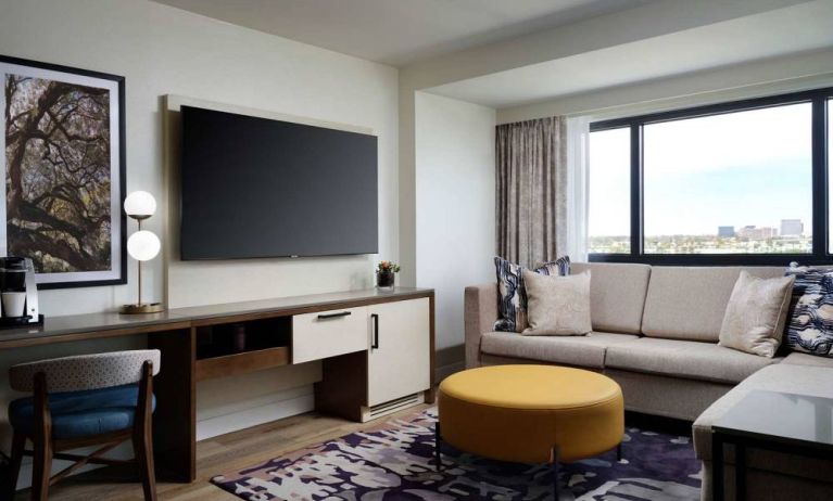 Sonesta Irvine guest room living area, including corner sofa, chair, window, and large, wall-mounted TV.