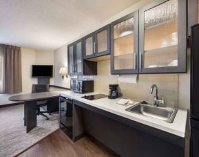 Sonesta Simply Suites Miami Airport Doral guest room kitchen, furnished with oven, microwave, and hob, plus a fridge-freezer, with table and chair nearby.