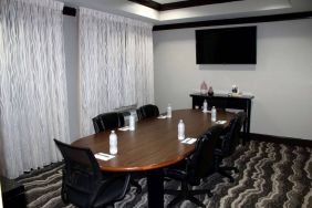 Hotel meeting room, including a wooden table and five leather chairs around it, two windows, and a large TV on the wall.