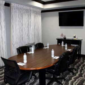 Hotel meeting room, including a wooden table and five leather chairs around it, two windows, and a large TV on the wall.