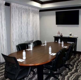 Hotel meeting room, including a wooden table and five leather chairs around it, two windows, and a large TV on the wall.