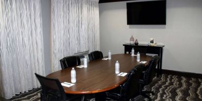 Hotel meeting room, including a wooden table and five leather chairs around it, two windows, and a large TV on the wall.