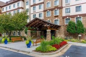 The hotel’s exterior has a pair of benches beside the covered entrance, with flowerbeds, bushes, and trees nearby.