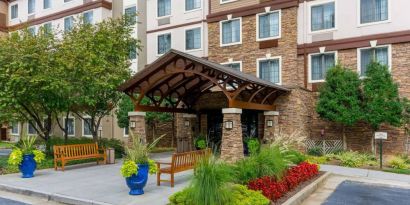 The hotel’s exterior has a pair of benches beside the covered entrance, with flowerbeds, bushes, and trees nearby.