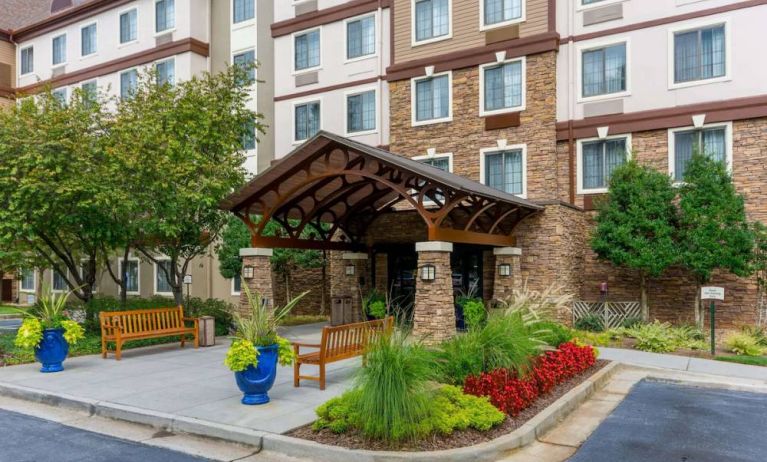 The hotel’s exterior has a pair of benches beside the covered entrance, with flowerbeds, bushes, and trees nearby.