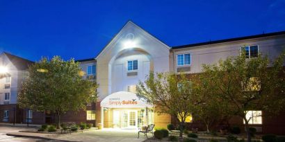 The hotel’s exterior has potted plants and benches by the front door, with trees and bushes nearby.