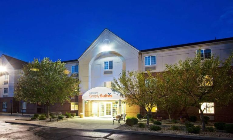 The hotel’s exterior has potted plants and benches by the front door, with trees and bushes nearby.