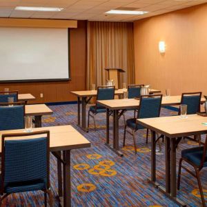 Hotel meeting room with seating for a dozen attendees and tables arranged in a classroom format facing a projector screen.