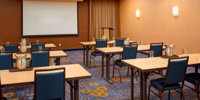 Hotel meeting room with seating for a dozen attendees and tables arranged in a classroom format facing a projector screen.