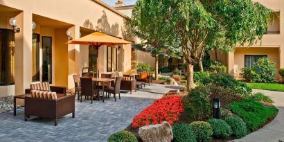 The hotel’s patio is furnished with a mix of tables and chairs, and sofa/armchair seating, next to flowerbeds and trees.