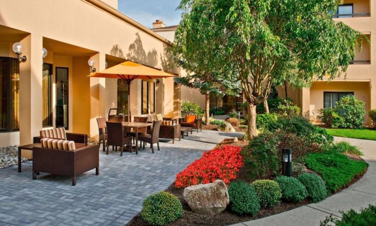 The hotel’s patio is furnished with a mix of tables and chairs, and sofa/armchair seating, next to flowerbeds and trees.