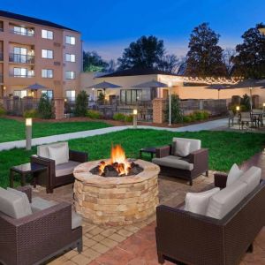 Sonesta Select Raleigh Durham Airport Morrisville’s fire pit has armchair and sofa seating, plus coffee tables, next to it, with shaded tables and chairs nearby.