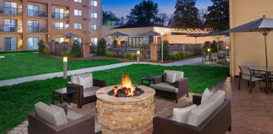 Sonesta Select Raleigh Durham Airport Morrisville’s fire pit has armchair and sofa seating, plus coffee tables, next to it, with shaded tables and chairs nearby.