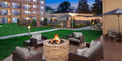 Sonesta Select Raleigh Durham Airport Morrisville’s fire pit has armchair and sofa seating, plus coffee tables, next to it, with shaded tables and chairs nearby.