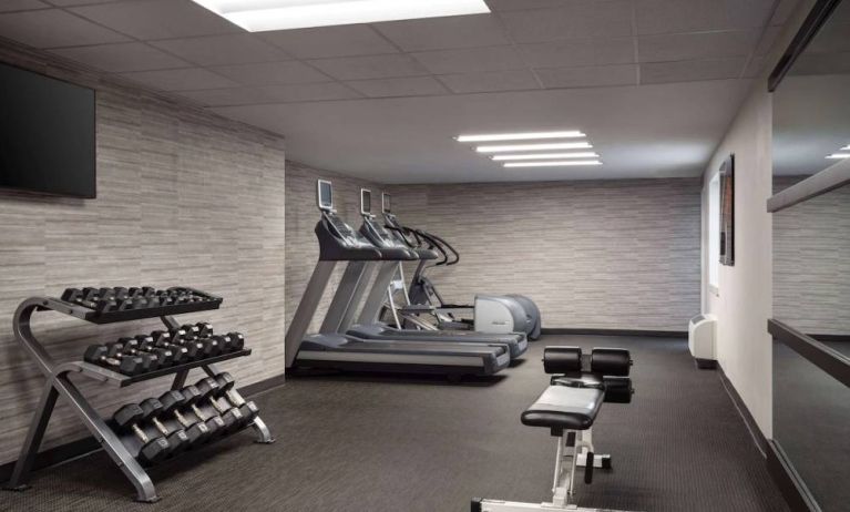 The fitness center at Sonesta Select Phoenix Camelback is equipped with free weights, a range of exercise machines, and a wall-mounted television.