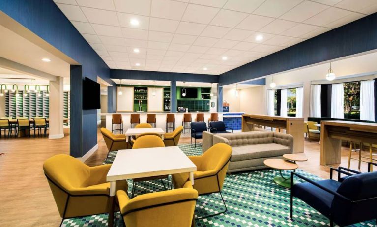 Sonesta Select Philadelphia Airport’s lobby lounge is furnished with tables and chairs, plus sofas and armchairs, and has a large, wall-mounted TV.