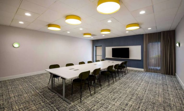 Hotel meeting room, with long table, a dozen chairs, spacious room, and a large TV mounted on the wall.