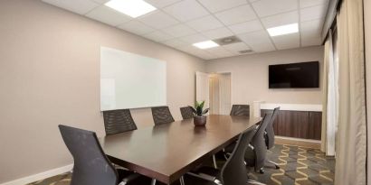 Hotel meeting room furnished with long wooden table, eight swivel chairs, a whiteboard, and a wall-mounted television.