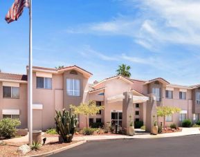 The hotel’s exterior has a fluttering USA flag flying from a flagpole and greenery including bushes, small trees, and a cactus.