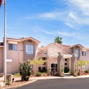 The hotel’s exterior has a fluttering USA flag flying from a flagpole and greenery including bushes, small trees, and a cactus.