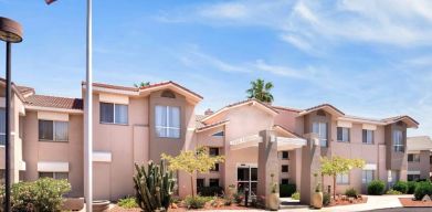 The hotel’s exterior has a fluttering USA flag flying from a flagpole and greenery including bushes, small trees, and a cactus.