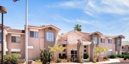 The hotel’s exterior has a fluttering USA flag flying from a flagpole and greenery including bushes, small trees, and a cactus.