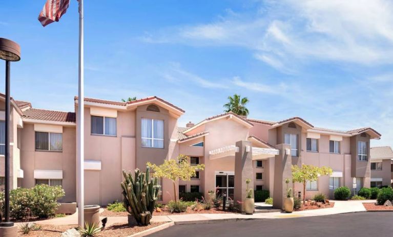 The hotel’s exterior has a fluttering USA flag flying from a flagpole and greenery including bushes, small trees, and a cactus.